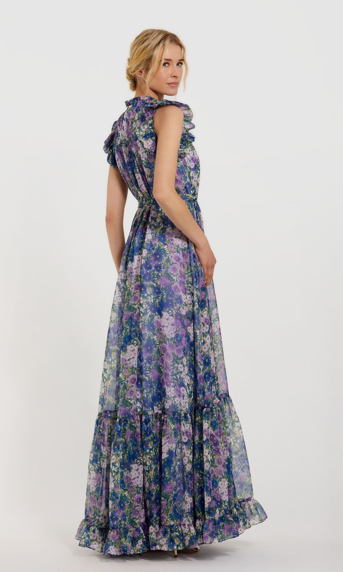 A woman models the Mac Duggal 56116 Lilac Floral-Print Formal Gown, a long sleeveless dress in purple, blue, and green tones, standing against a plain white background and looking over her shoulder.